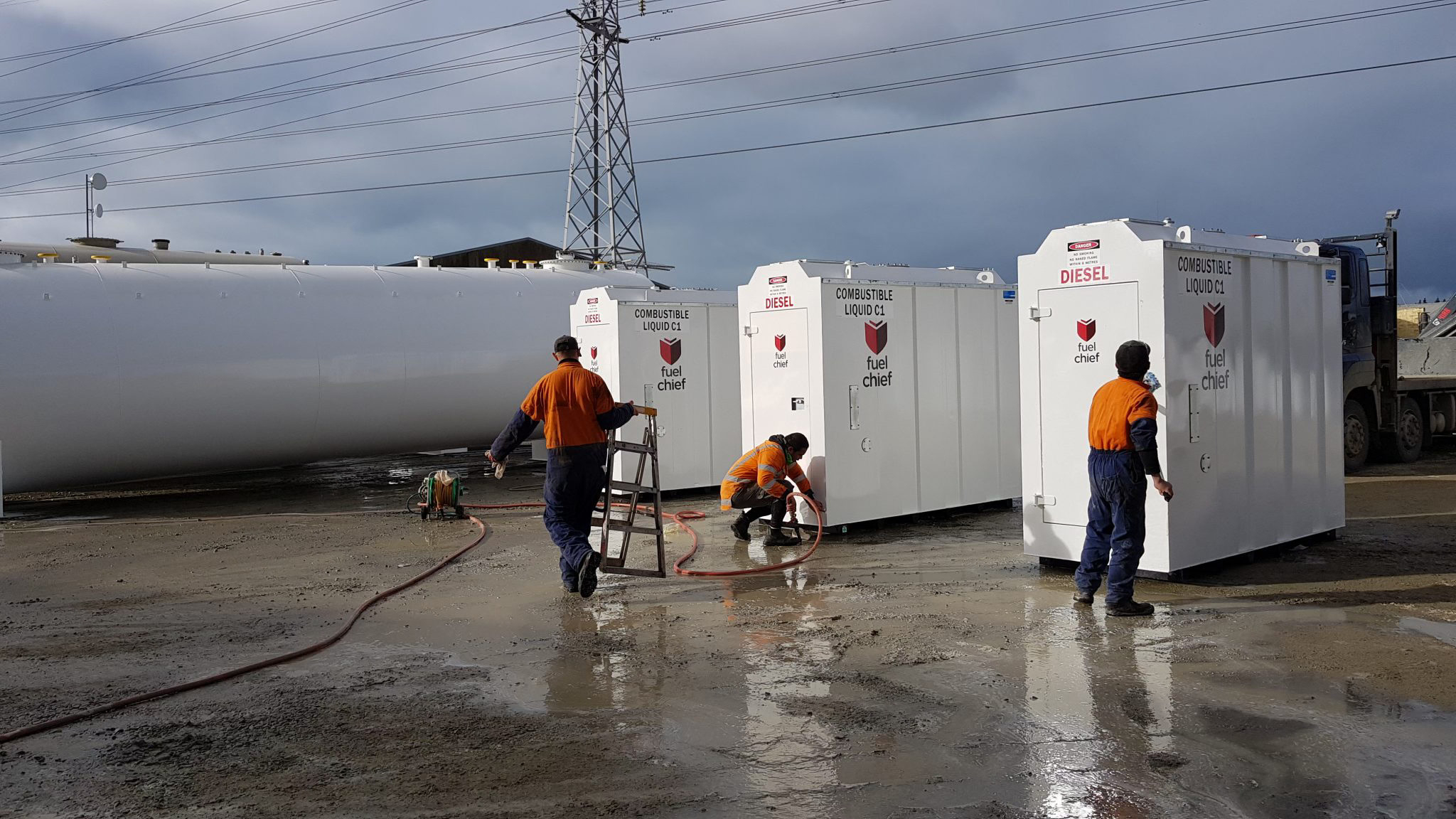 Workers in high-visibility gear preparing white Fuelchief tank enclosures in a wet yard