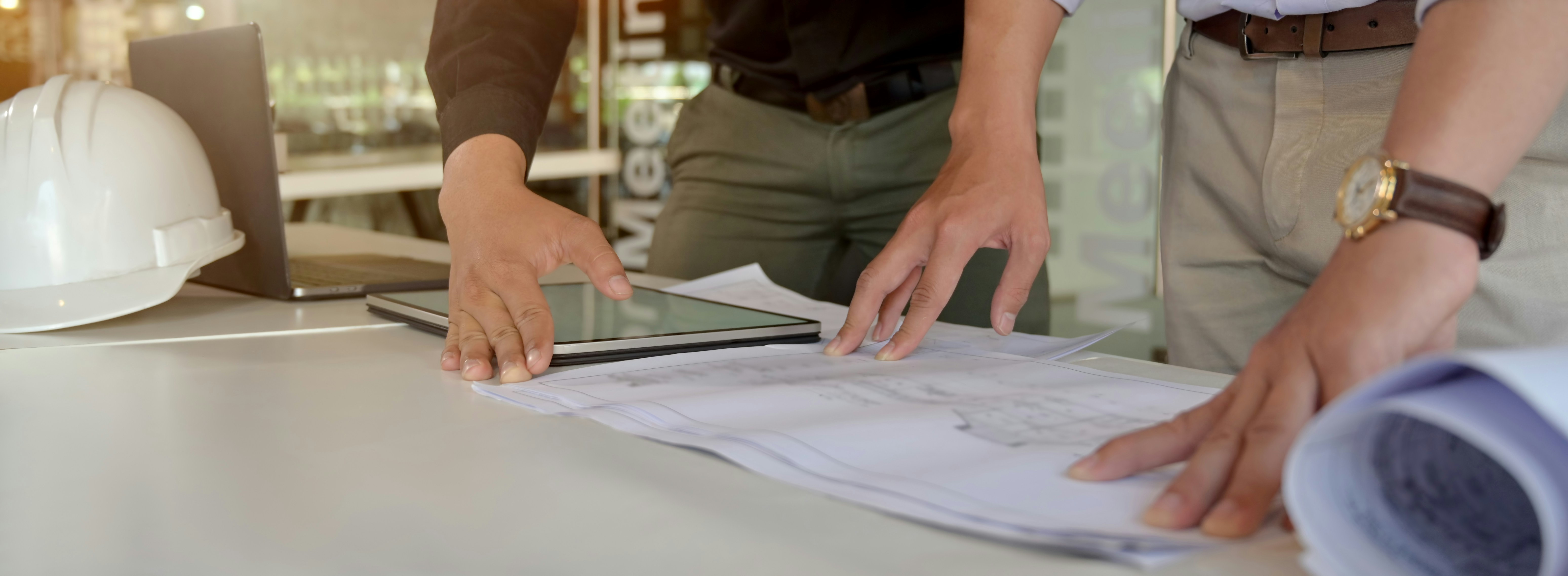 Two people reviewing drawings and a tablet on a desk beside a hard hat
