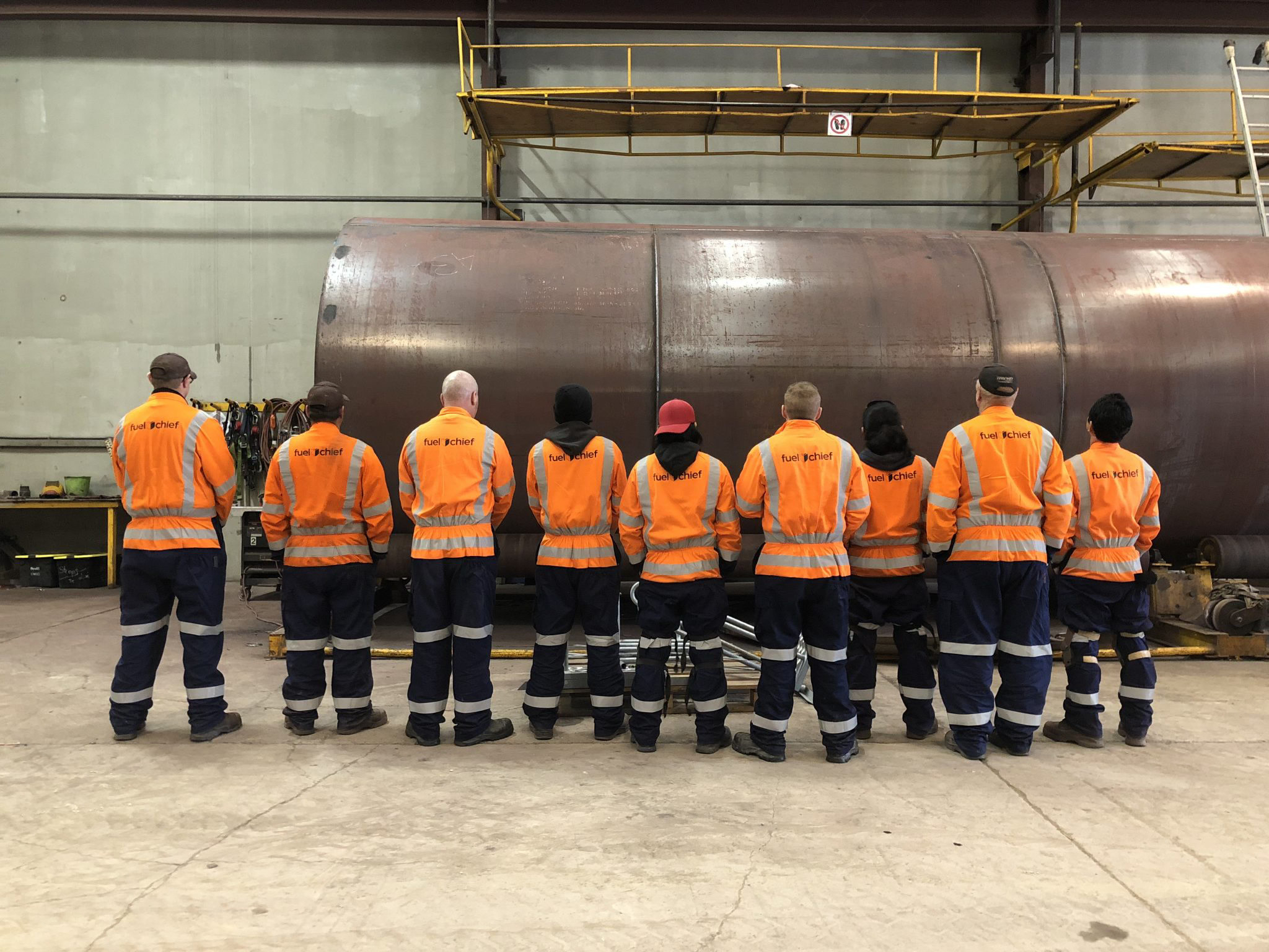 Fuelchief factory team in orange high-visibility jackets, seen from behind, facing a large steel tank