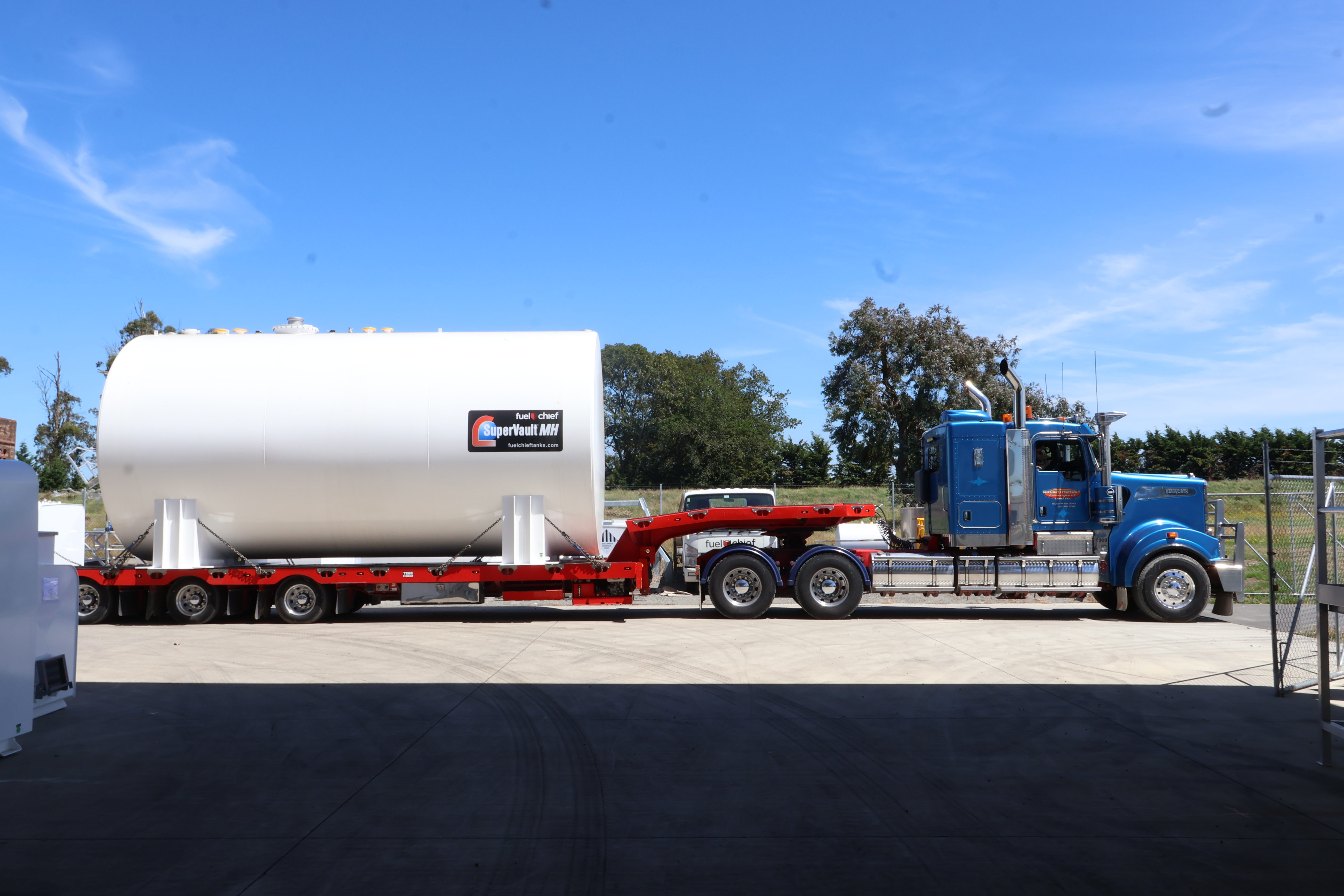 White fuel tank on a low-loader trailer pulled by a blue truck